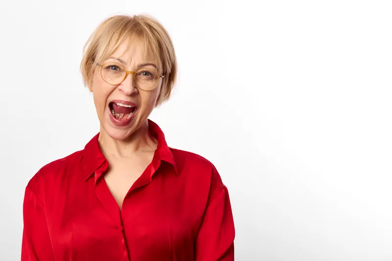 woman with glasses laughing in a studio portrait, red shirt, short blond hair, open mouth smile, bright white background, cheerful mood, friendly