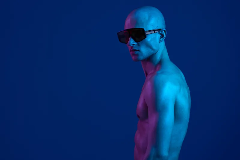 Bald man posing confidently in blue lighting with stylish sunglasses, showcasing a strong physique in a vibrant atmosphere. Perfect for fashion and advertising needs.