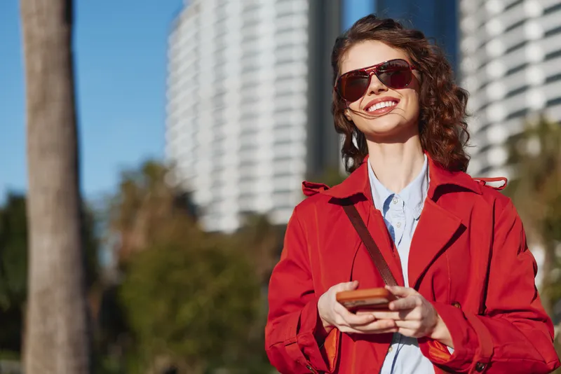 Young woman with curly hair wearing a red jacket and sunglasses smiles while holding a smartphone outdoors in a sunny urban setting with modern buildings in the background.