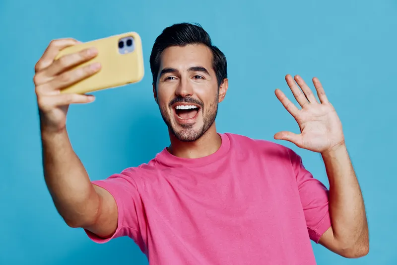 Smiling man in pink t-shirt waving hand while taking selfie with mobile phone, cheerful expression on his face, isolated on bright blue background. People lifestyle concept