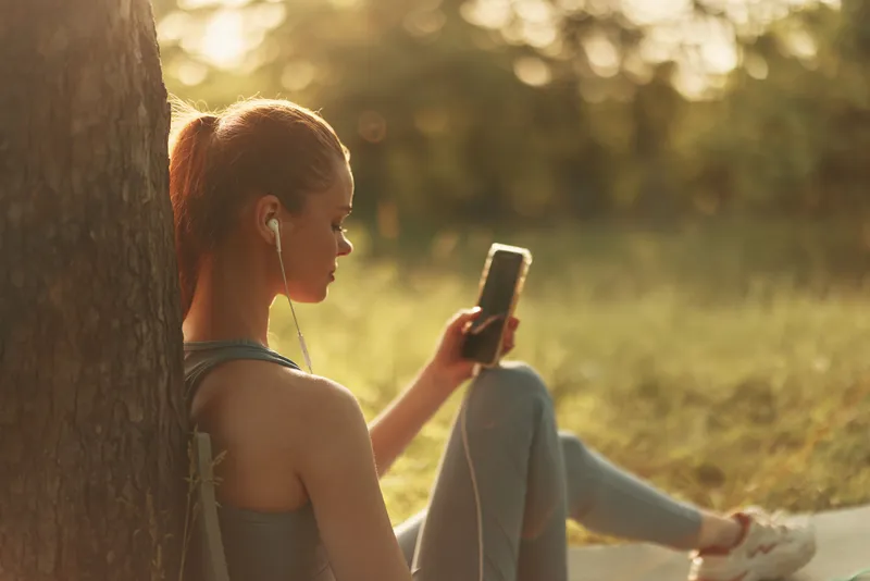Young woman enjoying music on her smartphone while sitting outdoors, dressed in casual athletic wear, expressing relaxation and calmness in a sunny park setting