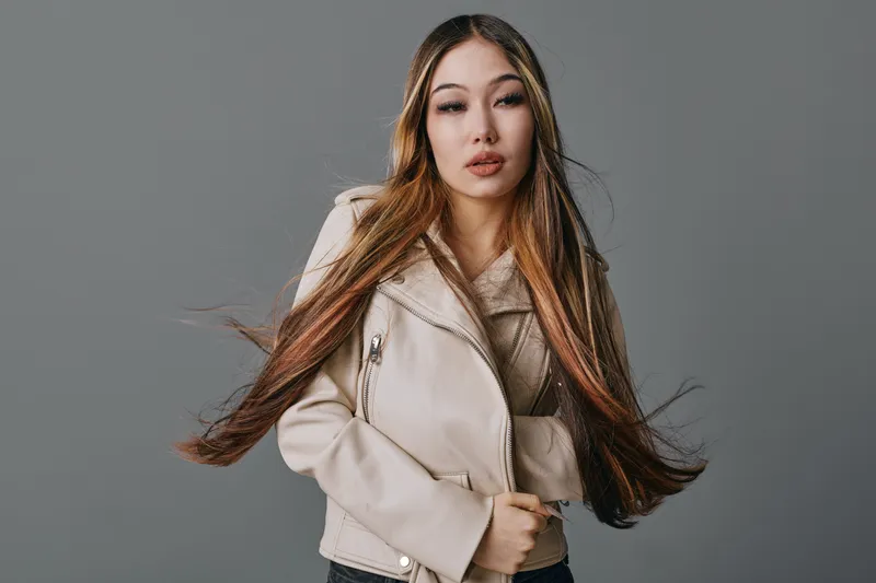 Young woman with long brown hair wearing beige jacket poses confidently against gray background in studio portrait. Attractive model with neutral expression and stylish outfit.