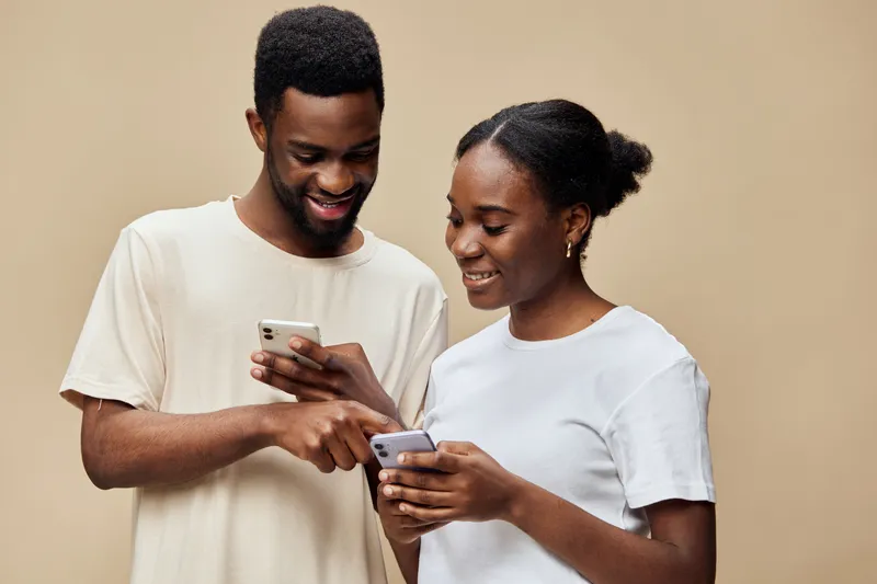 Young African couple smiling and using smartphones together, wearing casual light clothing against a neutral beige background, reflecting modern communication