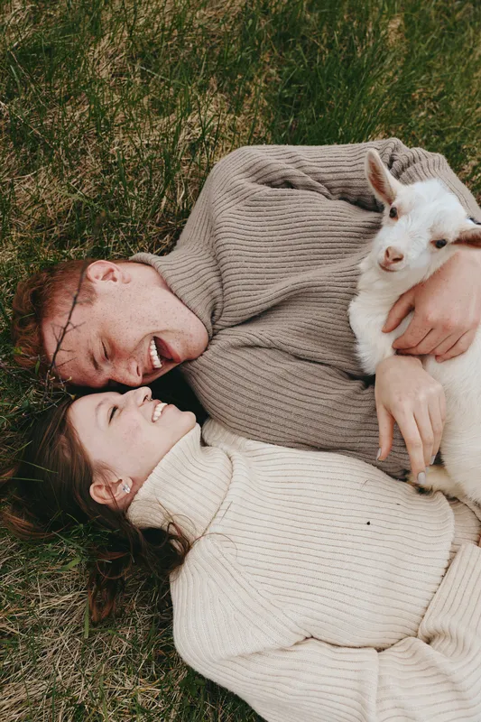 Warm lifestyle scene with a smiling couple on the grass holding a goat kid, cozy sweaters, and natural light creating a tender, carefree mood in a peaceful outdoor setting