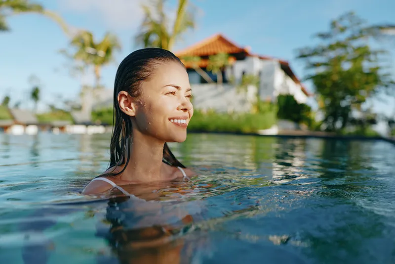 A smiling woman enjoys a refreshing pool swimming in sunlit water, with a tropical resort backdrop. This lifestyle shot conveys relaxation, leisure, happiness, and vacation vibes by the pool.