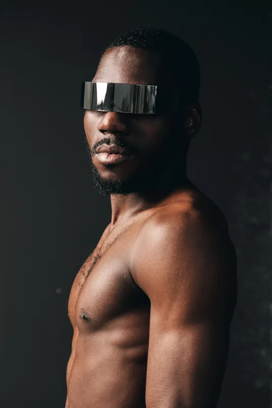 African man with dark skin and muscular body wearing futuristic silver visor in creative conceptual visual style. Portrait with strong lighting and modern artistic expression.