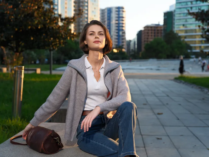 A stylish young woman sits on a park bench in an urban setting during golden hour. She wears a light blazer and jeans, exuding calm confidence in the city.