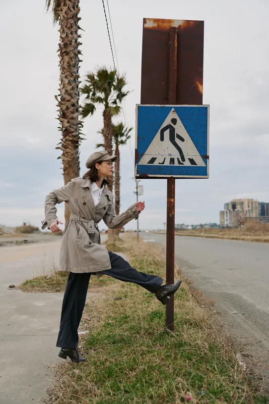 Woman in trench coat and hat stretches leg near rusty pedestrian crossing sign on roadside with palm trees and cloudy sky. Concept of urban lifestyle, fashion, and movement outdoors.
