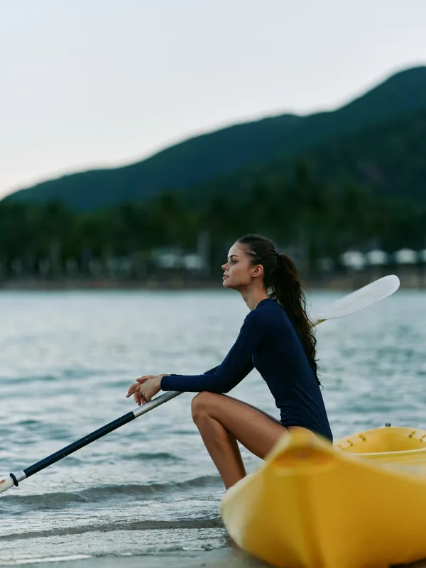 A focused kayaker sits on a yellow kayak at the shoreline, wearing a dark wetsuit, paddling with a long oar as gentle waves lap the water.