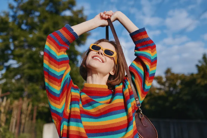 Woman in a colorful striped sweater and yellow sunglasses smiling outdoors; young adult joyful and carefree, raising a shoulder bag strap in a park scene for casual fashion travel lifestyle.