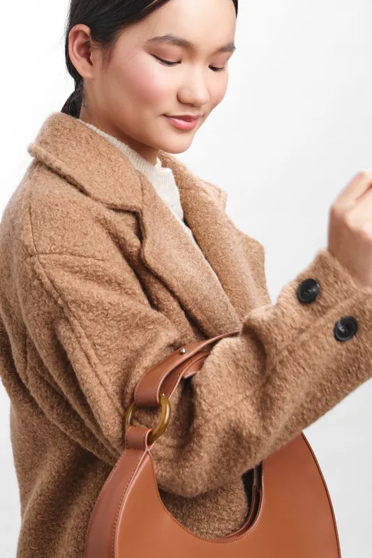 asian woman wearing a warm beige coat carries a brown handbag, looking downward with a gentle smile, exuding a calm, stylish urban mood suitable for fashion and lifestyle campaigns.