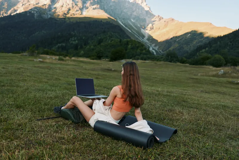 A lone woman sits on a mat in a meadow, using a laptop. She wears a bright top and shorts, embracing nature while working remotely and enjoying the scenic mountains.