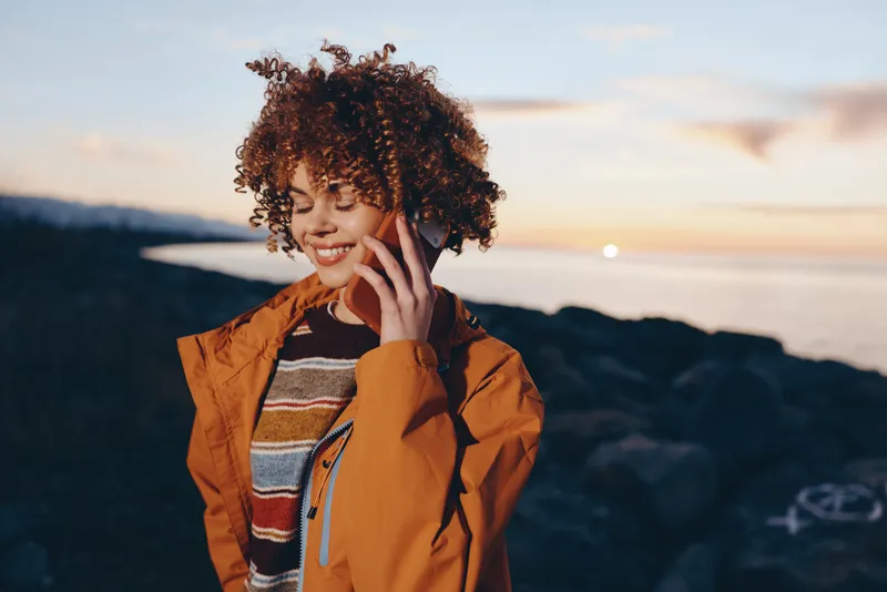 Cheerful woman lifestyle portrait outdoors at sunset, curly hair blowing softly, smiling and touching hair, wearing orange jacket, sea breeze and ocean waves, warm natural light