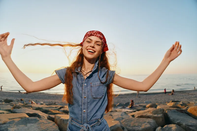 Modern boho-western denim street style woman joyfully waving hair on beach during golden hour, casual summer outfit with film color effect, lifestyle outdoor mood