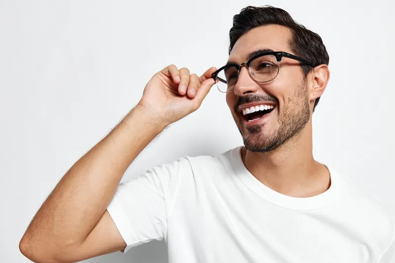 Full body smiling cheerful young man wearing glasses and a white t-shirt looking at the camera with a joyful expression isolated on a solid light background.