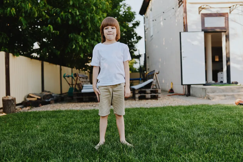Happy boy standing in a sunny backyard, wearing a white shirt and beige shorts. The bright green grass adds to the cheerful atmosphere of the scene.