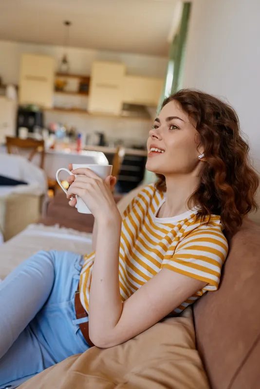 A woman relaxing at home on a sofa, holding a mug and wearing a striped shirt, looking up with a soft smile. A cozy, everyday moment in a bright living space.