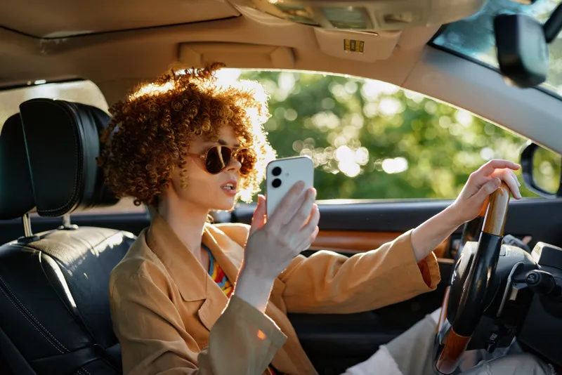 Curly haired woman driving a car while checking her phone, wearing sunglasses, daytime light, warm retro mood, casual chic style, road ahead, sense of independence and travel.