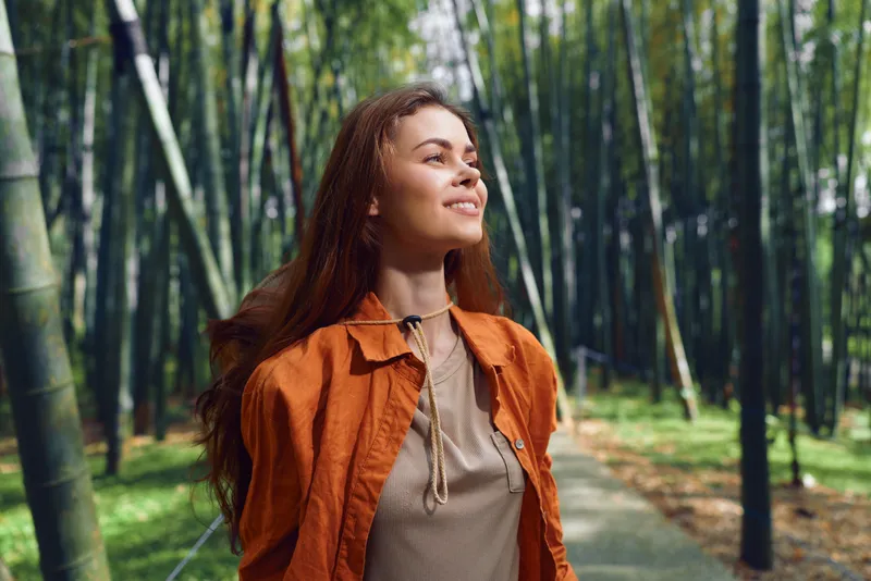 Woman portrait with smile in a bamboo forest enjoying nature outdoors on a shaded path. Young female in orange jacket looks upward, sunlight and calm atmosphere around her.