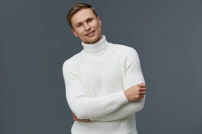 Smiling young caucasian man wear white knitted turtleneck sweater crossing arms looking at camera isolated on plain gray background studio portrait digital nomad concept
