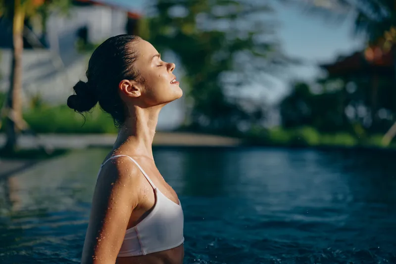 Young woman in white bikini enjoys sunlight with closed eyes standing in outdoor pool water during sunny day with blurred background and natural lifestyle mood.