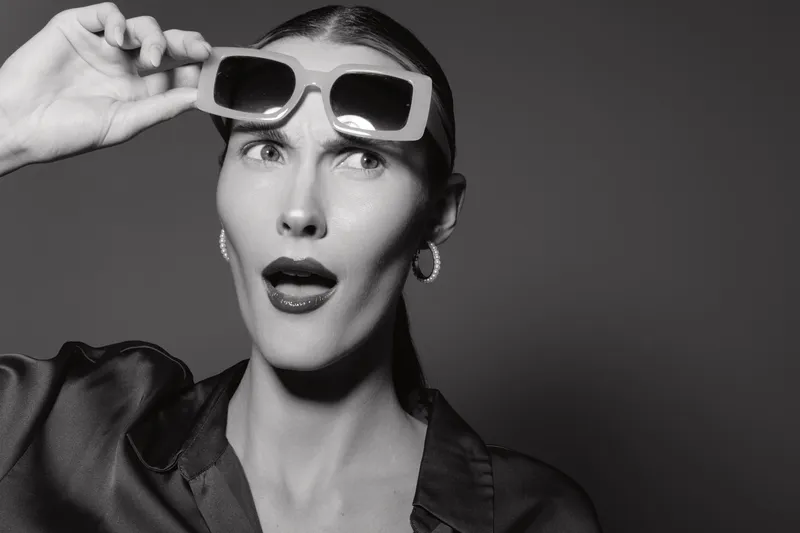 Caucasian woman with pale skin wearing sunglasses looks surprised, posing in studio with dark background. Fashion concept.