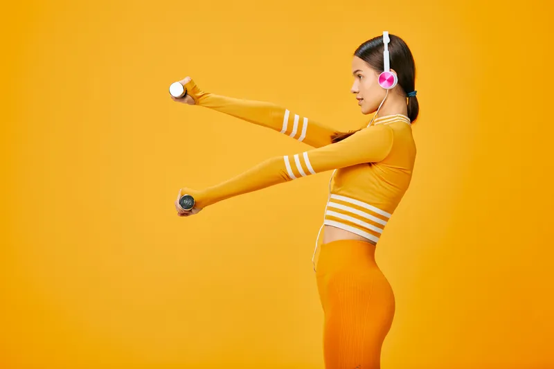 Fitness woman in vibrant orange activewear extends arms with light dumbbell in a bright studio, headset on, showcasing dynamic movement, energy and focus during workout.