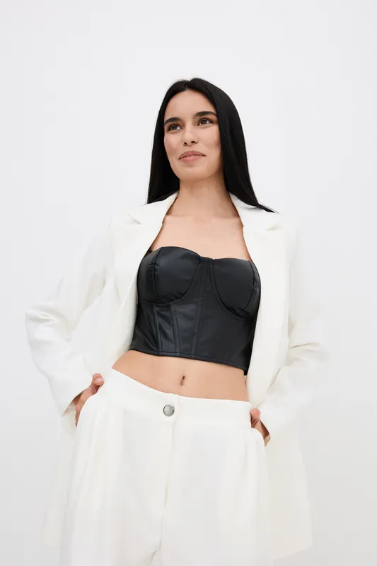 confident woman in white suit and black corset posing with hands in pockets against plain white background, showcasing modern fashion and minimalistic style.