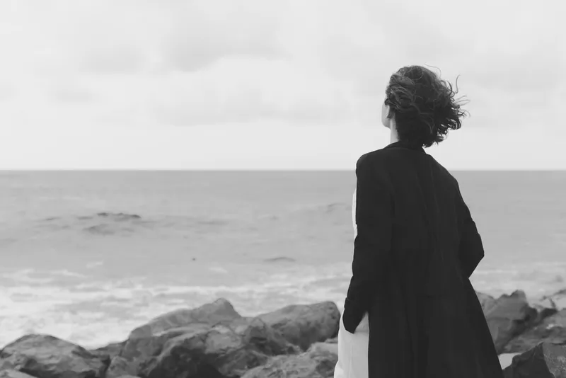 Silhouette of a woman standing on the coast in monochrome, facing the sea, wind in hair, calm seascape, distant rocks and thoughtful mood