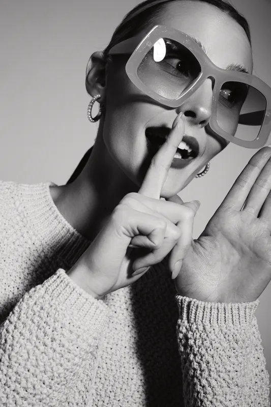 Smiling white woman with light skin wearing large sunglasses and knitted sweater makes hush gesture with finger near lips. Studio portrait in black and white with playful mood concept.