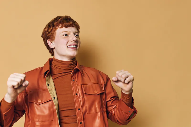 Young man with red hair wearing a brown leather jacket and turtleneck posing with happy smile and raised hands on beige background. Casual style and confident expression.