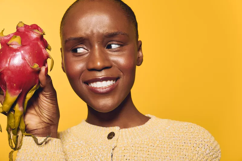 Young African woman with dark skin holds exotic dragon fruit near face, smiling slightly. Bright yellow background and soft lighting create fresh, healthy lifestyle concept.