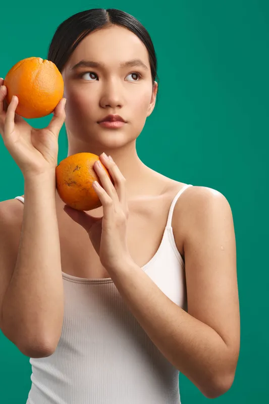 Asian woman with calm expression holds two oranges against a vivid green backdrop, wearing a white sleeveless top, creating a fresh, vibrant still life portrait.