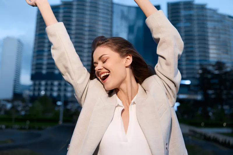 cheerful woman celebrating success outdoors in autumn wearing beige coat and white blouse with modern city skyscrapers background