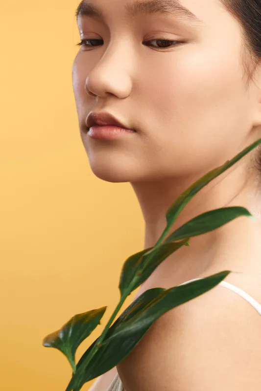 asian woman with a calm expression poses beside a green plant against a warm yellow background, soft lighting highlights smooth skin and a graceful profile, modern artistic portrait.