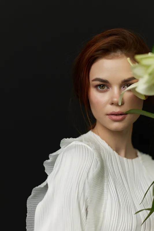 Elegant portrait of a female model with red hair, dressed in a white blouse, showcasing beauty and serenity against a dark backdrop with flowers