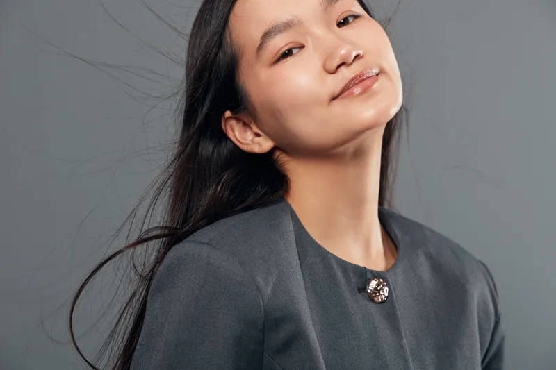 Young woman with long hair wearing dark grey blouse posing on grey background with confident expression and natural makeup, studio portrait with soft lighting and modern style.