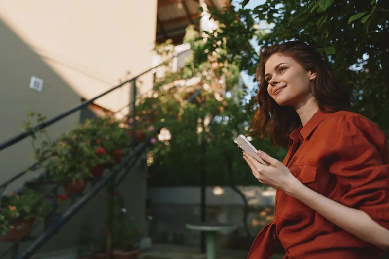 Young woman in a red shirt stands in a sunny garden, smiling while checking her phone, a casual lifestyle moment under warm light and lush greenery