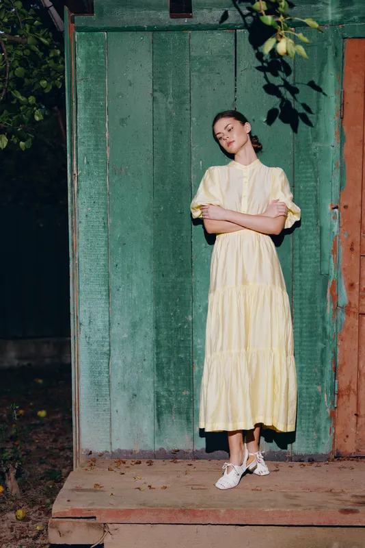 Woman in a pale yellow dress stands with arms crossed in front of a green wooden doorway, outdoors, exuding calm confidence and relaxed fashion.