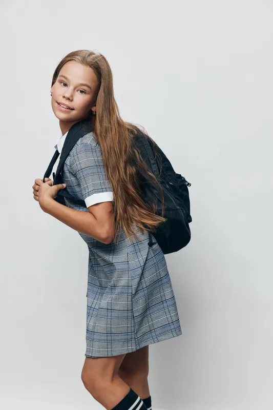 Schoolgirl with long hair wearing a backpack and plaid dress posing against a white background. Student ready for school or education with a smile and casual posture.
