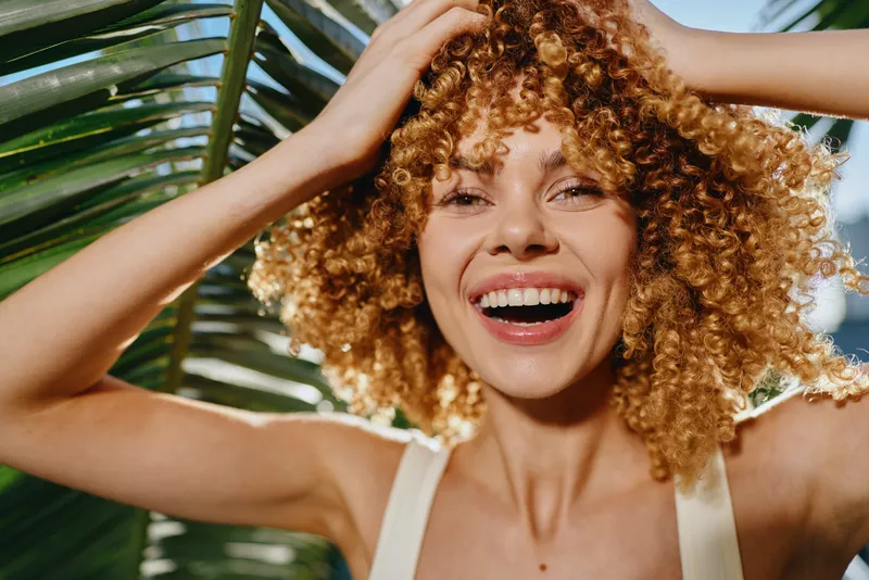 Smiling woman with curly hair joyfully posing outdoors near tropical palm leaves in bright natural sunlight, radiating happiness and summer vibes.