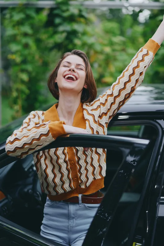 A joyful woman leans out of a car door, laughing with a bright expression against a lush outdoor backdrop.