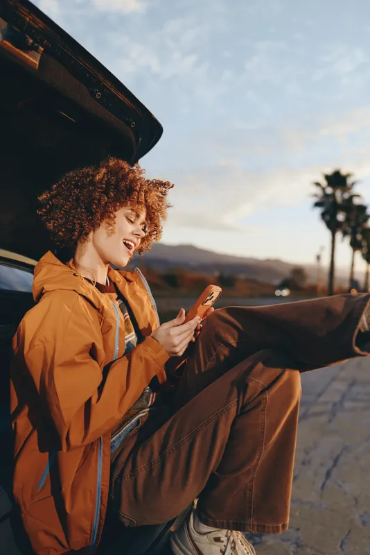 Woman lifestyle, curly haired woman in orange jacket sits by car trunk, using smartphone outdoors at sunset, mountain road background, relaxed travel mood, casual fashion