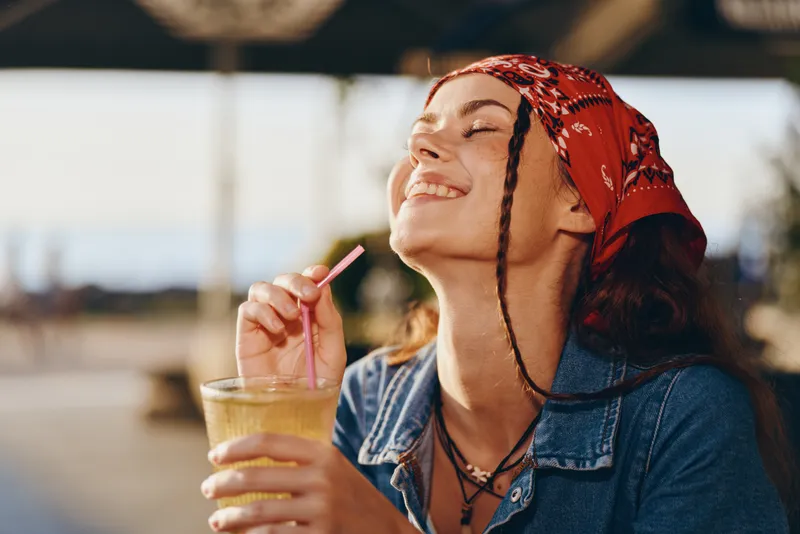 smiling woman enjoying fresh drink outdoors with joyful expression, casual denim jacket, red headscarf, warm sunlight, relaxed mood, summer vibe