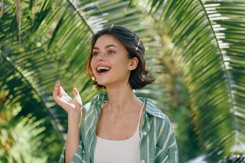 Young woman outdoors, smiling and looking up, wearing a light green shirt over a white top, with lush tropical palm leaves in the bright background.