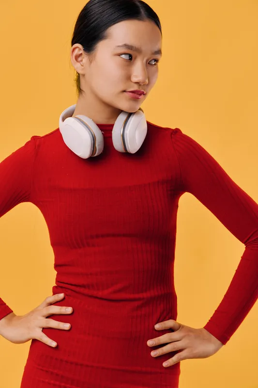 Young woman in red turtleneck with white headphones around neck standing confidently with hands on hips against orange background. Casual style and focused expression.