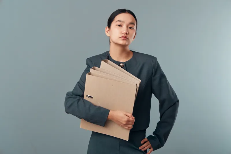 Young woman in professional suit holding cardboard folders with documents on plain background for office work, businesswoman, organization and serious expression in portrait.