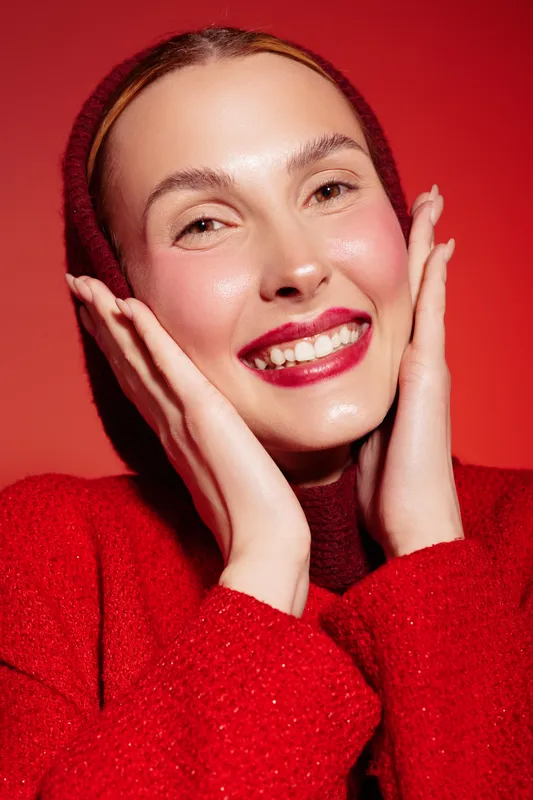 Smiling woman with red lipstick and rosy cheeks wears red sweater and headscarf, posing with hands near face on red background. Concept of beauty, fashion, and joyful expression.
