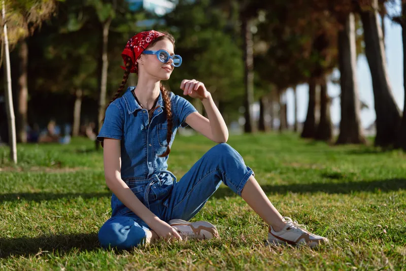 lifestyle woman in modern boho-western denim street style sitting outdoors on green grass with a red bandana and blue sunglasses, film color effect enhancing colors
