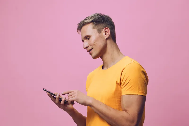 A full body young man wearing bright yellow t-shirt, smiling and looking at his phone, standing against a plain pink background in studio portrait. People lifestyle concept
